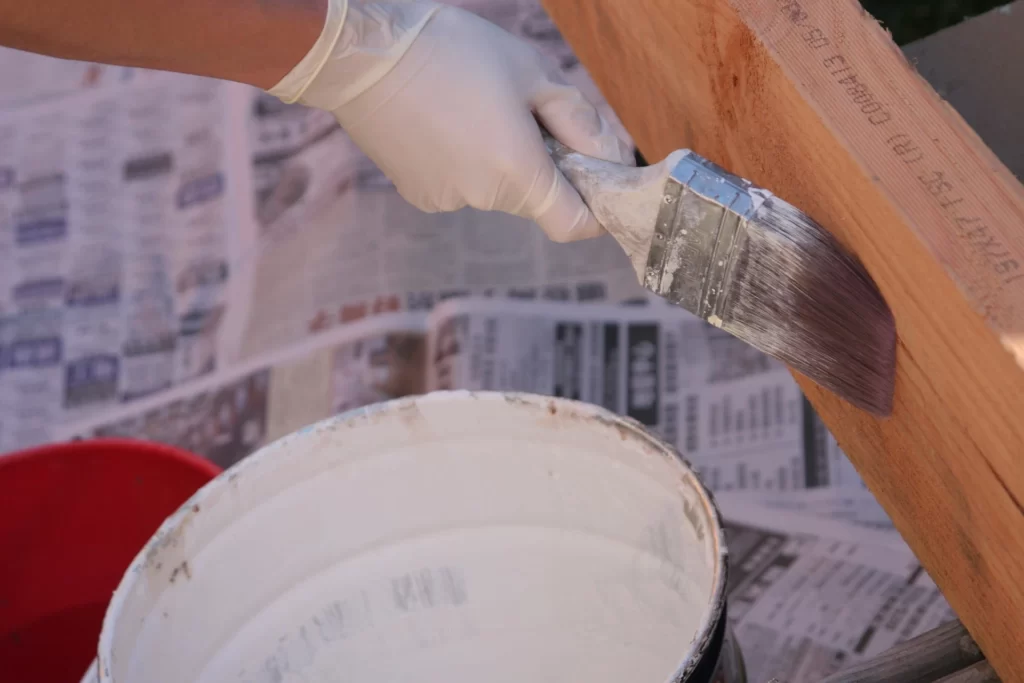 person using a paintbrush to apply a brown stain to a wooden deck, with a pile of tools and the deck partially covered in plastic sheeting.